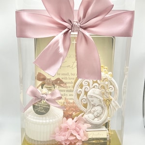 May include: A clear gift box with a pink satin ribbon and bow. The box contains a white candle with a gold lid, a small white figurine of the Virgin Mary holding a baby, a gold chain with a cross, and a pink floral arrangement. The candle has a tag that says "Thank you for being my Godmother Love Avelia".