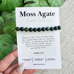 Könnte beinhalten: Ein Moss-Achat-Kristallarmband mit dunkelgrünen Perlen wird auf einer weißen Karte gezeigt. Auf der Karte steht "Moss Agate Crystal Jewellery" und enthält Text über die Eigenschaften des Steins. Grüne Blätter sind im Hintergrund.