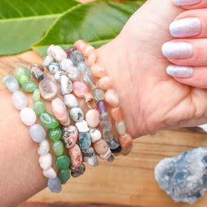 Op de afbeelding: Een stapel van acht armbanden gemaakt van verschillende halfedelstenen, waaronder groene jade, roze rozenkwarts en witte opaal. De armbanden zijn gestapeld op de pols van een persoon.