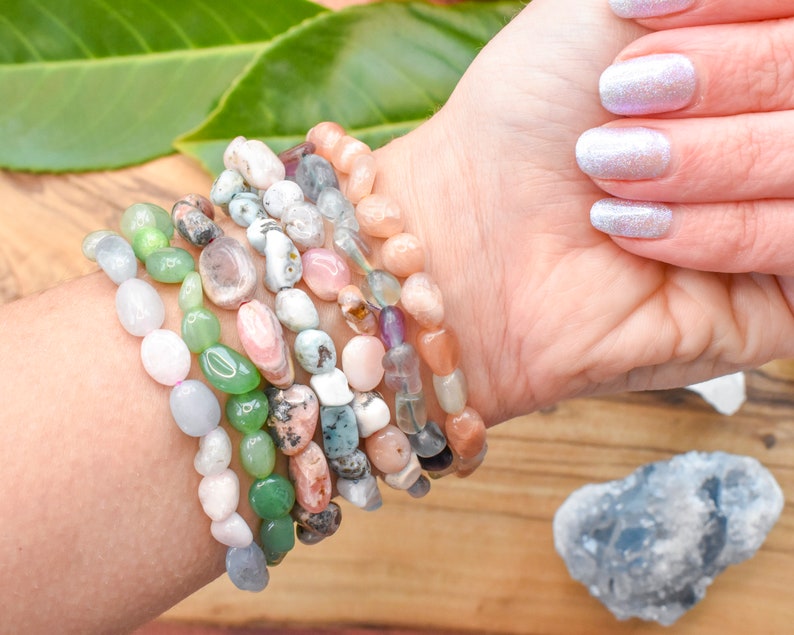 Op de afbeelding: Een stapel van acht armbanden gemaakt van verschillende halfedelstenen, waaronder groene jade, roze rozenkwarts en witte opaal. De armbanden zijn gestapeld op de pols van een persoon.