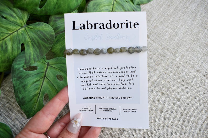 May include: A Labradorite crystal bead bracelet on a white card with text. The card says "Labradorite Crystal Jewellery" and describes the stone's properties. The bracelet features gray and green beads.