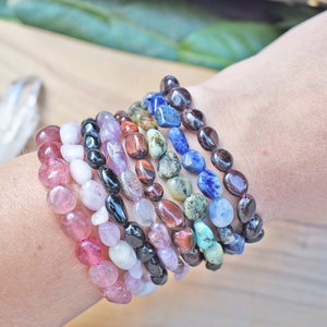 May include: A stack of colorful gemstone bracelets, including pink, white, black, purple, red, green, blue, and brown. The bracelets are made of smooth, rounded stones.