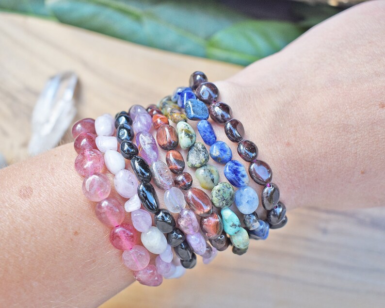 May include: A stack of colorful gemstone bracelets, including pink, white, black, purple, red, green, blue, and brown. The bracelets are made of smooth, rounded stones.