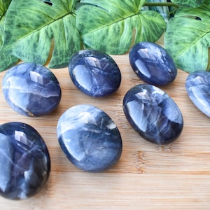 May include: Seven polished, oval-shaped blue stones with white veining are arranged on a wooden surface. The stones have a glossy finish and reflect light. Green leaves are in the background.
