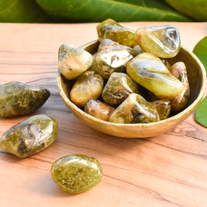 May include: A selection of polished green gemstones in a small, round, wooden bowl. The stones are various shades of green and brown. The bowl is on a wooden surface, with green leaves in the background. The gemstones are likely measured in grams.