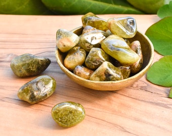 May include: A collection of tumbled green gemstones in a small, round, wooden bowl. The stones have a smooth, polished appearance and vary in shades of green and brown. The bowl sits on a wooden surface, with green leaves in the background.