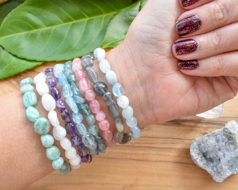 May include: A selection of gemstone bracelets in shades of turquoise, white, purple, blue, pink, and grey. The bracelets, made of smooth, rounded beads, are shown on a wrist.