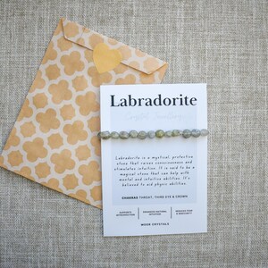 May include: A labradorite crystal bracelet is displayed on a card with the word "Labradorite" printed on it. The card is next to a patterned envelope with a heart-shaped sticker. The bracelet is made of small, round, grey-green stones.