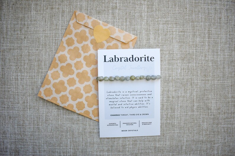 May include: A labradorite crystal bracelet is displayed on a card with the word "Labradorite" printed on it. The card is next to a patterned envelope with a heart-shaped sticker. The bracelet is made of small, round, grey-green stones.