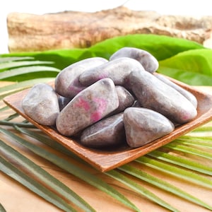 May include: A wooden square bowl filled with smooth, grey stones with pink markings. The stones are arranged on a wooden surface with green leaves and a piece of wood in the background.