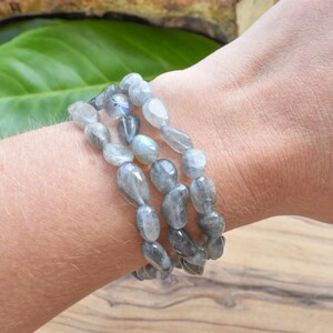 May include: Three strands of grey labradorite gemstone bracelets. The beads are oval-shaped and have a polished, smooth surface. The bracelets are worn stacked on a wrist.