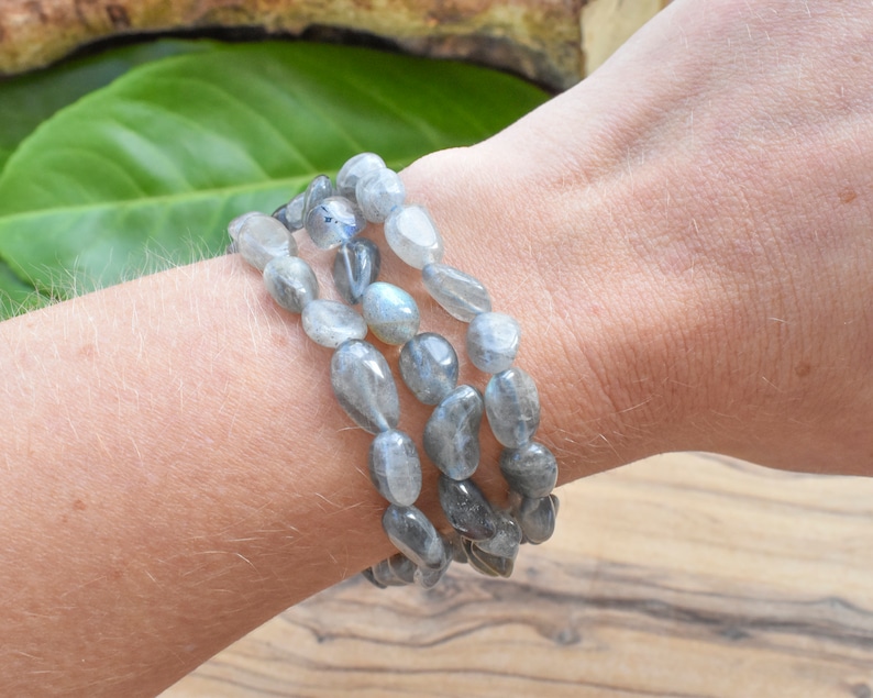 May include: Three strands of grey labradorite gemstone bracelets. The beads are oval-shaped and have a polished, smooth surface. The bracelets are worn stacked on a wrist.