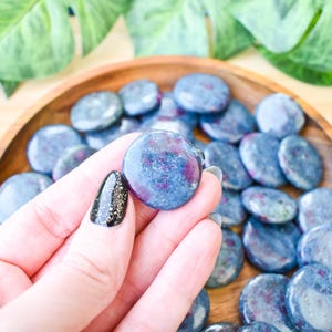 May include: A selection of polished, circular gemstones in blue and purple hues, presented in a wooden bowl. The gemstones are held by a hand with black and gold glitter nail polish. Green leaves are in the background.