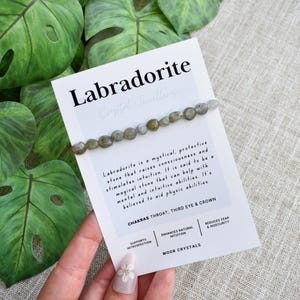 May include: A Labradorite crystal bracelet on a white card with text, next to large green leaves. The card reads "Labradorite" and describes the stone's properties. The bracelet features small, round, grey-green beads.