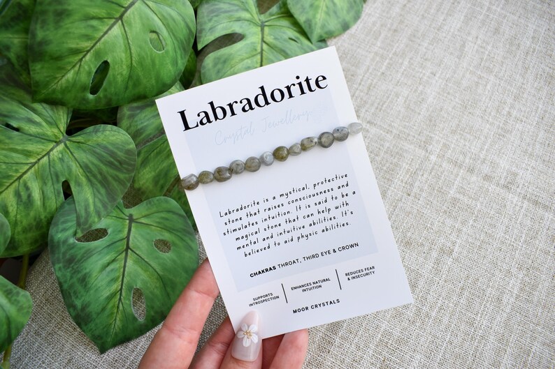 May include: A Labradorite crystal bracelet on a white card with text, next to large green leaves. The card reads "Labradorite" and describes the stone's properties. The bracelet features small, round, grey-green beads.