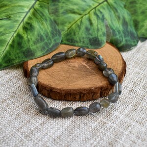 May include: A bracelet made of grey-green, oval-shaped beads. It sits on a small, round wooden slice, with green leaves in the background. The bracelet is a piece of jewellery.