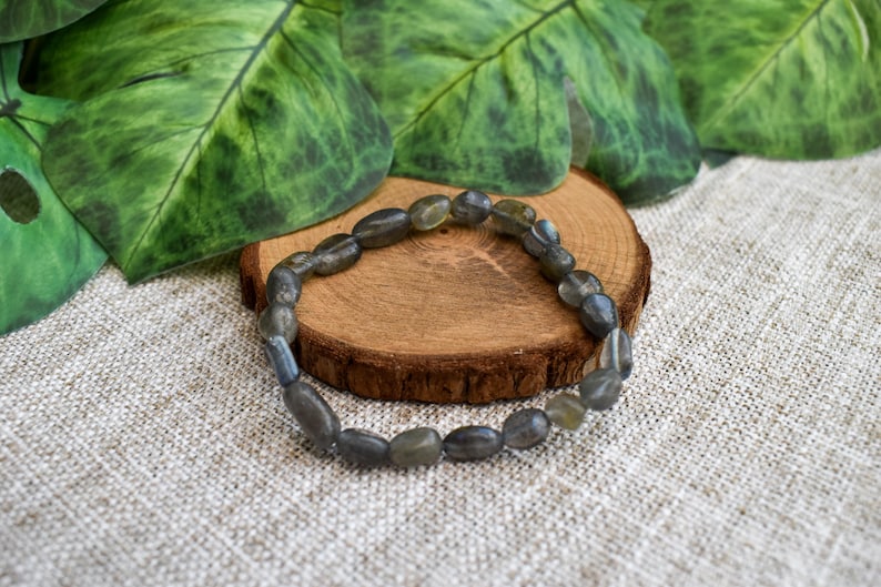 May include: A bracelet made of grey-green, oval-shaped beads. It sits on a small, round wooden slice, with green leaves in the background. The bracelet is a piece of jewellery.