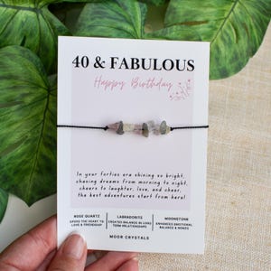 May include: A white card with the text "40 & FABULOUS Happy Birthday" and a bracelet with pink and grey stones. The card also includes text about chasing dreams and the benefits of rose quartz, labradorite, and moonstone.