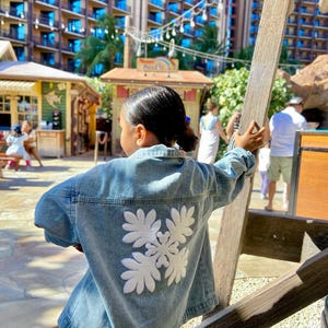 May include: A light blue denim jacket with a white floral design on the back. The jacket is worn by a person standing near a wooden structure. The background includes buildings and other people.