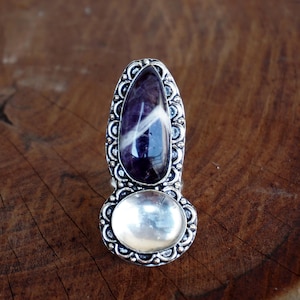 May include: A silver ring featuring two gemstones. The top stone is a large, teardrop-shaped amethyst with white streaks. The bottom stone is a smaller, round, clear gemstone. The ring has an ornate, filigree-style setting.