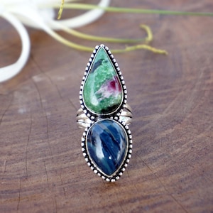 May include: A silver ring featuring two stacked, pear-shaped cabochon gemstones. The top stone is mottled green with a patch of deep pink, while the bottom stone is a dark, striated blue. The metal setting has a beaded edge and a split band design.