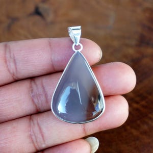 May include: A teardrop-shaped pendant with a polished, gray gemstone set in a silver-colored metal frame. The pendant is suspended from a silver bail. The gemstone has a glossy surface.