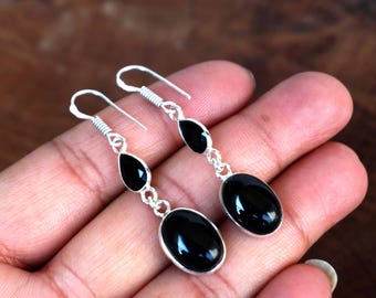 May include: A pair of dangle earrings featuring three black onyx stones on each earring. The stones are oval and teardrop-shaped, set in a silver metal. The earrings are displayed against a warm, brown background.