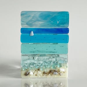 May include: A layered glass sculpture depicting a seascape. The bottom layer is white with brown and white glass pieces representing sand and shells. The next layer is light blue with white swirls representing waves. The next layer is a darker blue, and the top layer is a light blue with white swirls representing clouds. Two small white glass pieces represent birds flying in the sky.