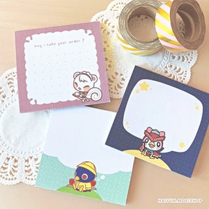 May include: Three square notepads with cartoon animal illustrations. One notepad is pink with the text "may I take your order?". Another is blue with stars, and the third is green. Decorative washi tape rolls are in the background.