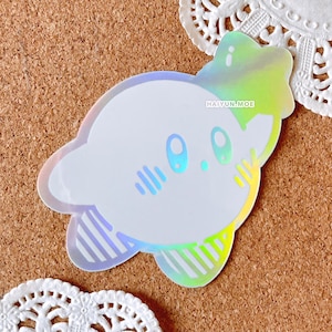 May include: A holographic sticker of a white Kirby character with a pink blush and closed eyes. The sticker has a rainbow iridescent finish and the text "HAIYUN.MOE" is printed on the sticker.