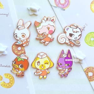 May include: Six different Animal Crossing character enamel pins. Each pin features a different character wearing a white and red uniform with a white hat. The characters include a squirrel, a dog, a cat, a rabbit, a duck, and a horse. The pins are all rose gold in color.