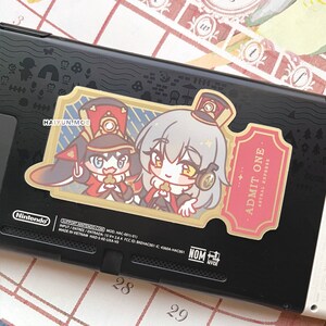 May include: A sticker featuring two anime characters in military uniforms. The characters are standing in front of a gold ticket with the text "ADMIT ONE ASTRAL EXPRESS".