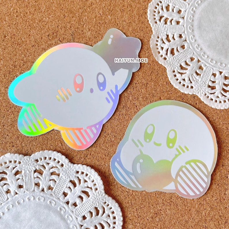 May include: Two iridescent holographic stickers of a cartoon character with large eyes and a pink blush. The character is holding a heart in one sticker and a star in the other. The stickers are on a corkboard background.