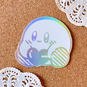 May include: A holographic sticker featuring a white cartoon character with a pink blush and closed eyes. The character is holding a striped object in each hand. The sticker has the text "HAI YUN.MOE" printed on it.