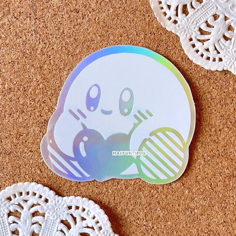 May include: A holographic sticker featuring a white cartoon character with a pink blush and closed eyes. The character is holding a striped object in each hand. The sticker has the text "HAI YUN.MOE" printed on it.