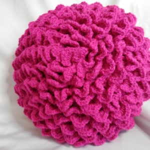 May include: A bright pink crocheted pillow with a ruffled texture. The pillow is round and has a soft, fluffy appearance.
