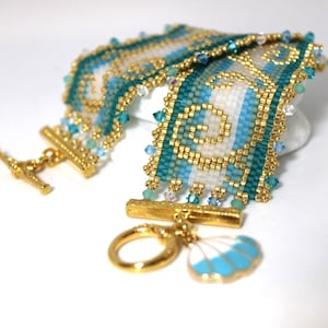 May include: A beaded bracelet with a gold clasp and a seashell charm. The bracelet features a pattern of white, teal, and gold beads. Small blue and clear crystals are also incorporated into the design. The bracelet is approximately 7 inches long.