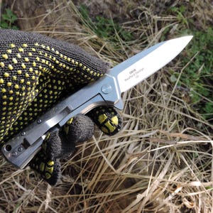 May include: A silver folding knife with a sharp blade is held in a black glove with yellow dots. The knife has a clip and the text "0808, Rexford Design, D2" is visible on the blade. The background is blurred with grass and foliage.