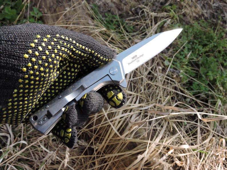 May include: A silver folding knife with a sharp blade is held in a black glove with yellow dots. The knife has a clip and the text "0808, Rexford Design, D2" is visible on the blade. The background is blurred with grass and foliage.