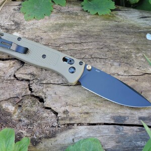 Könnte beinhalten: Ein Klappmesser mit schwarzer Klinge und beigefarbenem Griff. Das Messer hat einen schwarzen Taschenclip und einen goldfarbenen Drehpunkt. Die Klinge ist etwa 8,9 cm lang.