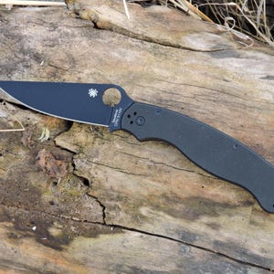 Può includere: Un coltello pieghevole nero con un manico curvo e una lama parzialmente seghettata. La lama ha un foro circolare vicino al dorso e il marchio è visibile. Il coltello è appoggiato su una superficie di legno stagionato.