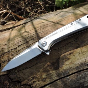 May include: A silver folding knife with a sharp blade, resting on a weathered wooden surface. The knife has a sleek, metallic handle with the text "ZERO TOLERANCE" etched on the side. The blade is open, showcasing its sharp edge.