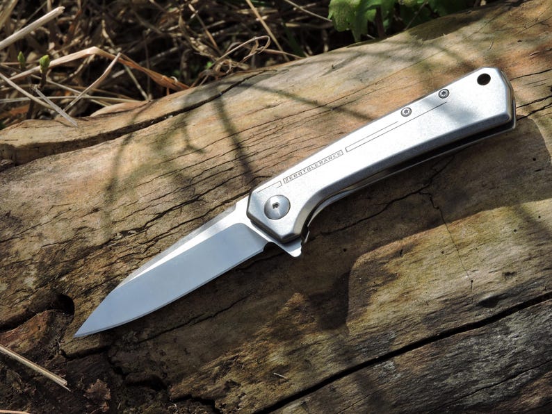 May include: A silver folding knife with a sharp blade, resting on a weathered wooden surface. The knife has a sleek, metallic handle with the text "ZERO TOLERANCE" etched on the side. The blade is open, showcasing its sharp edge.