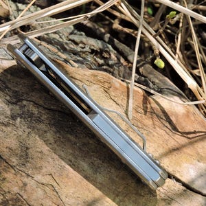 May include: A silver folding pocket knife with a clip, resting on a weathered wooden surface. The knife is closed, showing its sleek, metallic side. The background includes dry grass and twigs, suggesting an outdoor setting.