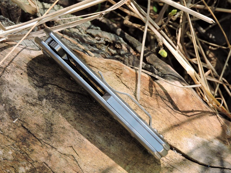 May include: A silver folding pocket knife with a clip, resting on a weathered wooden surface. The knife is closed, showing its sleek, metallic side. The background includes dry grass and twigs, suggesting an outdoor setting.