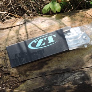 May include: A white ceramic mug with a black handle and the text "But First, Coffee" in a bold, black font. The mug is filled with a dark liquid, likely coffee, and sits on a light-colored surface.