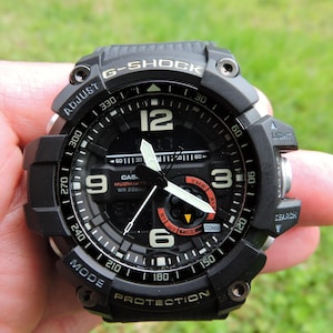 May include: A black Casio G-Shock Mudmaster watch featuring an analog and digital display with white hour markers, luminous hands, and text including G-Shock, Mudmaster, WR 20BAR, Adjust, Light, Search, Mode, and Protection on the bezel.