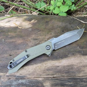May include: A folding pocket knife with an olive green handle and a grey metallic blade. The handle features a black pocket clip with the Kershaw brand name printed on it. The blade is marked with the text 1575 E48 8Cr13MoV near the pivot.