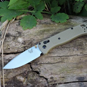 May include: A tan folding knife with a silver blade, featuring a black butterfly logo and black hardware. The handle has a textured grip. The knife is open and resting on a weathered wooden surface, with green foliage in the background.