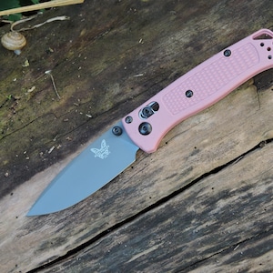 May include: A folding pocket knife with a matte grey drop-point blade featuring a butterfly logo and a textured light pink handle. The handle includes black hardware, a pivot screw, and a lanyard hole at the end of the grip for secure carrying.
